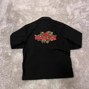 Svart overshirt från Hollister - Svart overshirt från Hollister med stor broderad logga i rött och gult på ryggen. Skjortan har klassisk krage, knappar framtill och två bröstfickor med lock. Materialet är mjukt och känns som bomull. Perfekt för dig som vill ha en chill och snygg look.
