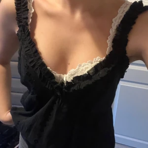 Svart linnetopp med spetsdetaljer - Säljer en svart linnetopp med breda axelband och v-ringning. Toppen har fina volangdetaljer längs halsringningen och vit spetskant som ger en romantisk vibe. Materialet känns lätt och luftigt, perfekt för varma dagar. Snygg att styla med jeans eller kjol.