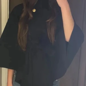 Svart capejacka med guldknapp - Snygg svart capejacka med hög krage och en dekorativ guldknapp vid halsen. Jackan har vid passform och vida ärmar som ger en cool och elegant siluett. Perfekt för dig som vill sticka ut med en stilren look.