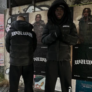 Svart pufferjacka från WUWA XS - Svart pufferjacka från WUWA med stor vit logga på ryggen och mindre logga på bröstet. Jackan har huva, dragkedja framtill och två sidofickor med dragkedja. Fylld med syntetmaterial och har en loose passform, perfekt för kalla dagar.