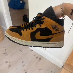 Nike Air Jordan 1 Mid svart/brun mocka - Säljer ett par Nike Air Jordan 1 Mid i svart och brun mocka med klassisk vinge-logga på sidan. Skorna har svarta snören, vit sula och ikonisk swoosh. Perfekta för dig som vill sticka ut med en snygg färgkombo och streetstyle-vibe.