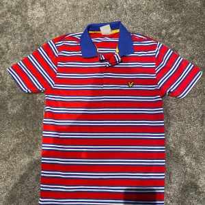 Randig piké från Lyle & Scott Golf - Färgstark randig pikétröja från Lyle & Scott Golf i rött, blått och vitt med blå krage och broderad gul logga på bröstet. Tillverkad i 100% polyester, perfekt för golf eller chill dagar. Klassisk passform och korta ärmar.