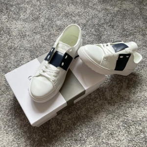 Valentino Garavani Open sneakers vit/blå - Snygga Valentino Garavani Open sneakers i vitt skinn med bred marinblå rem och silvriga nitar på sidorna. Klassisk rund tå, vita snören och platt sula. Perfekta för dig som vill ha en lyxig och stilren sneaker med exklusiva detaljer.