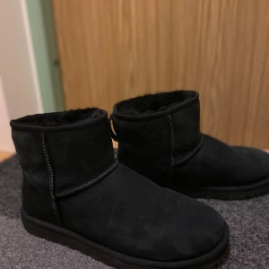 Svarta mockaboots med fårfoder - Mysiga svarta boots i mocka med mjukt fårfoder på insidan. Skorna har en rund tå, platt sula och klassisk söm längs sidorna. Perfekta för kalla dagar när du vill hålla fötterna varma och sköna.