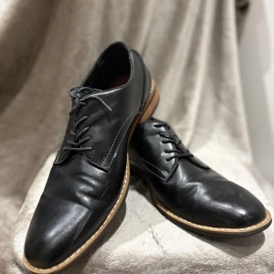 Svarta skinnskor med snörning - Klassiska svarta skinnskor från Cavalet med snörning och låg klack. Skorna har rund tå, kontrasterande brun sula och stilren design som passar till både jeans och kostym. Perfekta för dig som vill ha en tidlös och clean look.