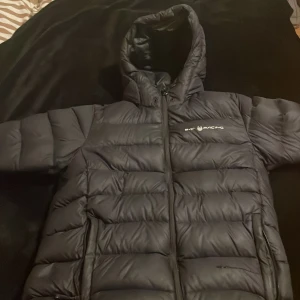 Svart pufferjacka från BAT RACING - Säljer en svart pufferjacka från BAT RACING med huva och dragkedja framtill. Jackan är vadderad och har en quiltad look med breda kanaler. Perfekt för kalla dagar och har en clean, sportig vibe.