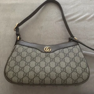 axelväska med GG-mönster - Snygg axelväska  med klassiskt GG-mönster i grått och svart. Väskan har guldfärgad logodetalj, justerbar axelrem och dragkedja upptill. Perfekt storlek för det viktigaste och en ikonisk design som sticker ut.