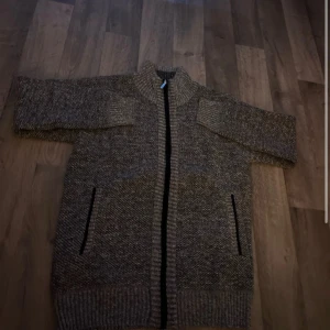 Grå stickad kofta med dragkedja - Mysig gråmelerad stickad kofta med hel dragkedja framtill och två fickor. Insidan har ett rutigt foder i brunt, svart, blått och vitt som ger extra värme. Koftan har ribbade muddar och hög krage, perfekt för kyliga dagar. 