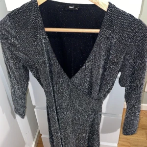 Glittrig svart klänning Filippa K - Supersnygg svart glittrig klänning från Filippa K med v-ringning och trekvartsärm. Klänningen har en omlott-look och är perfekt för dig som vill sticka ut med stil. Materialet är mjukt och följsamt med massor av glittereffekt.