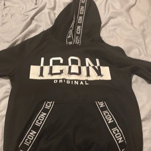 Svart hoodie ICON Original med huva - Svart hoodie från ICON Original med stor vit broderad text på bröstet och vita detaljer på huvan och fickan. Tröjan har huva med snörning och känguruficka framtill. Perfekt för dig som gillar streetwear och tydliga loggor.