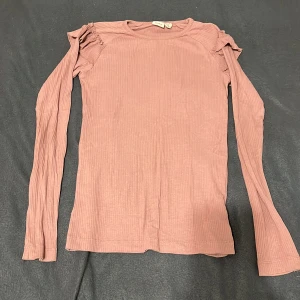 Rosa ribbad långärmad tröja Name It - Säljer en ljusrosa långärmad tröja från Name It i ribbad bomull. Tröjan har volangdetaljer vid axlarna som ger en söt och trendig touch. Perfekt för dig som gillar mjuka material och snygga detaljer.