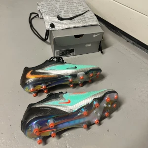 Nike Phantom GX Elite Ag fotbollsskor - Nike Phantom GX Elite Ag fotbollsskor i svart, turkos och silver med orange detaljer. Köpta på unisport för 3099 kr. Skorna har snörning, rund tå och platt sula med fasta dobbar. Ovandelen är i syntetmaterial och designen är riktigt modern med metallic-effekt och tydliga Nike-loggor på sidorna. Har används 1 gång och säljer pga att de är för små. Skopåse samt Nike box medföljer på köpet. Bara och höra av er vid funderingar eller frågor!