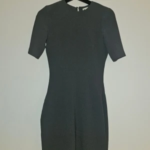 Mellan mörkgrå klänning med dragkedja - En stilren grå klänning med korta ärmar och rund hals. Klänningen har en figurnära passform och en diskret dragkedja baktill som gör den enkel att ta på. Perfekt för dig som gillar minimalistisk och klassisk stil. Öppen rygg och en liten knapp i naken att fästa med.