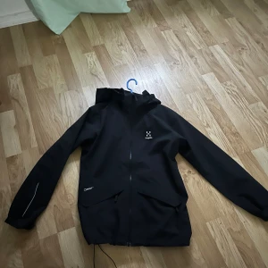 Svart regnjacka från Haglöfs med huva - Svart regnjacka från Haglöfs med huva och hel dragkedja framtill. Jackan har två stora fickor med lock, reflekterande detalj på ärmen och meshfoder på insidan. Perfekt för blöta dagar och outdoor-äventyr.