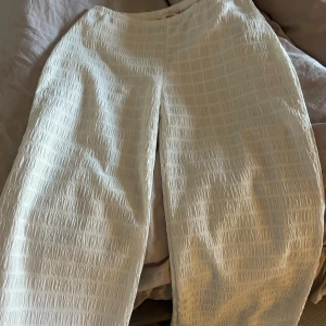 Vita wide byxor från Nelly XXS - Säljer ett par vita byxor från Nelly i storlek XXS. De har ett snyggt strukturerat tyg med rutmönster och är höga i midjan. Modellen är wide och ger en chill och trendig look. Perfekta för dig som gillar stilrena och bekväma plagg.