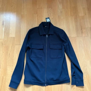 Mörkblå overshirt från Dressmann - Snygg mörkblå overshirt från Dressmann med hel dragkedja framtill och två stora fickor med lock på bröstet. Jackan har klassisk krage och långa ärmar med knapp vid mudden. Perfekt för lager-på-lager och stilren look. Ej använd