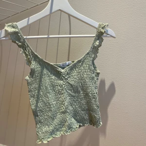 Ljusgrön smockad linnetopp Stradivarius - Supersöt ljusgrön linnetopp från Stradivarius med smockad struktur och volangkant. Axelbanden är spetsprydda och toppen har en v-ringning framtill. Perfekt för varma dagar och enkel att styla med jeans eller kjol.