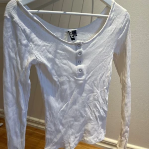 Vit ribbad långärmad topp från PIECES - Säljer en vit långärmad topp från PIECES med ribbad struktur och fyra silvriga knappar framtill. Toppen har en bred, rundad halsringning och är figurnära i modellen. Perfekt för dig som gillar enkel och clean stil.