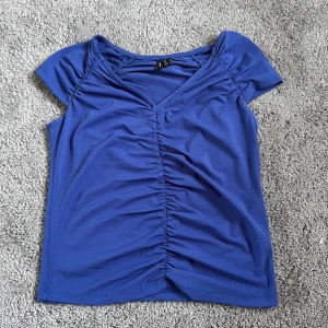 Blå topp från Vero Moda - Snygg blå topp från Vero Moda i storlek S. Modellen har korta ärmar, v-ringad hals och cool rynkdetalj längs mitten framtill. Materialet är mjukt och stretchigt, perfekt för dig som gillar en bekväm men stilren look.