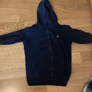 Marinblå hoodie från Lyle & Scott - Snygg marinblå hoodie från Lyle & Scott med dragkedja framtill och klassisk huva. Tröjan har en gul broderad logga på bröstet och praktisk känguruficka. Perfekt för chill dagar eller när du vill ha en clean streetstyle.