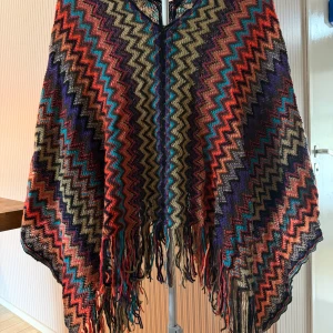 Färgglad stickad poncho med fransar - Poncho i stickat material med zigzag-mönster i lila, röd, beige, blå och orange. Snygga fransar längs nederkanten och loose passform. Perfekt för dig som gillar boho-vibe och vill ha något unikt och färgstarkt i garderoben.