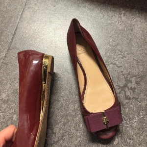 Vinröda pumps från Tory Burch - Snygga vinröda pumps från Tory Burch med öppen tå och låg klack i guld. Skorna har en stor rosett framtill med guldfärgad logga och är tillverkade i glansigt skinn. Perfekt för dig som gillar eleganta detaljer och vill sticka ut.