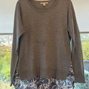 Grå tröja med blommigt tyg från Esprit - Snygg grå långärmad tröja från Esprit med rund hals och ribbade muddar. Nedtill syns ett blommigt tyg i vitt, blått och rosa som ger en cool lager-på-lager känsla. Perfekt för dig som gillar att sticka ut med detaljer och mönster.