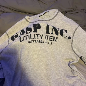 Ljusgrå långärmad tröja från GASP - Säljer en ljusgrå långärmad tröja från GASP med svart tryck både fram och bak. Tröjan har synliga sömmar och är tillverkad i mjuk bomull, perfekt för en avslappnad och sportig stil.