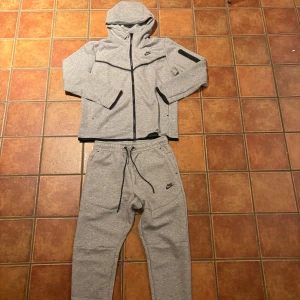 Grå Nike techflecce set - Nike tracksuit i ljusgrått med huva och hel dragkedja på tröjan. Byxorna har snörning i midjan och svarta detaljer vid fickorna. Både jacka och byxa har Nike-logga. Mjukt material, perfekt för chill eller träning. Snygg och sportig look.