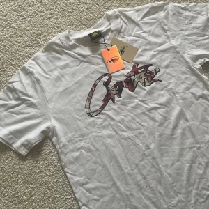 Vit t-shirt från Grizzly med tryck - Snygg vit t-shirt från Grizzly med stort grafiskt tryck i rött och svart på bröstet. Klassisk passform med rund hals och korta ärmar. Perfekt för dig som gillar streetwear och vill sticka ut med en cool logga framtill.