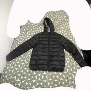 Svart pufferjacka med huva - Svart pufferjacka  med huva och dragkedja framtill. Jackan har quiltad design och är fodrad i grått. Perfekt för kalla dagar och har en clean, modern look som funkar till det mesta. Passformen är normal och jackan har långa ärmar.