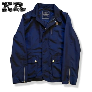 Morris Feild Jacket - Snygg marinblå vindjacka från Morris med klassisk krage, guldfärgade detaljer och dragkedjor vid ärmslut. Jackan har två stora fickor med knappar och ett Morris-emblem på bröstet. Insidan har diskret prickigt foder för extra stilpoäng.