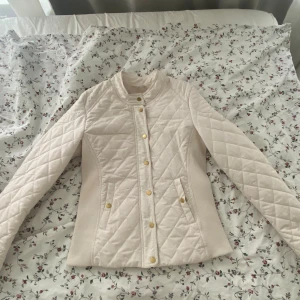 Quiltad vit jacka Holly & Whyte - Snygg quiltad jacka från Holly & Whyte i vit färg med guldiga knappar och dragkedja. Jackan har lång ärm, ståkrage och figurnära passform. Materialet är polyester och jackan har ribbade partier i sidorna för extra komfort.