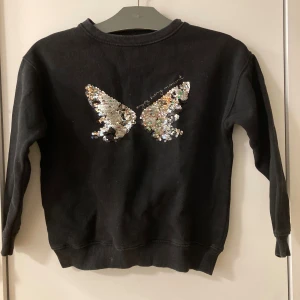 Svart tröja med blädderpaljettee - Svart college med lång ärm och rund hals. På bröstet finns ett snyggt paljettmotiv i silver, motiv av fjäril. I använt skick,  men fortfarande bra! Det enda är att den svarta färger i tröjan inte är så skarp svart. Annars hel och ren! 