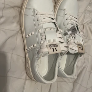 Valentino Rockstud vita sneakers - Säljer ett par vita Valentino Garavani sneakers med platt sula Skorna har ikoniska pyramidnitar på sidorna och tungan ganska nya använt bara 3 fester men kännde att skorna var för små för mig perfekt för dig som har storlek 42-43 går o förhandla vill sälja snabbt