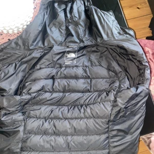 Svart pufferjacka från The North Face - Säljer en svart pufferjacka från The North Face i storlek S. Jackan har huva, dragkedja framtill och är quiltad för extra värme. Perfekt för kyliga dagar och har en clean, sportig look. Loggan syns tydligt på bröstet. Fråga gärna om fler bilder eller info! Ny pris ca 2500