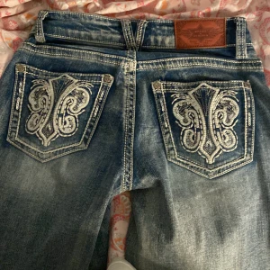 Stars & Stripes blå jeans med broderi OBS upplagda - Säljer ett par blå jeans från Stars & Stripes med snyggt slitna detaljer, low waist och  stora broderade bakfickor med silverfärgat mönster. Klassisk femficksmodell och bootcut ben. Perfekta för dig som vill sticka ut med coola detaljer.