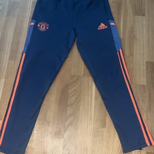 Manchester United träningsbyxor Adidas - Mörkblå träningsbyxor från Adidas med Manchester United-logga och orangea detaljer längs sidorna. Byxorna har smal passform, elastisk midja och är tillverkade i ett syntetmaterial som andas. Perfekta för fotbollsträning eller chill.