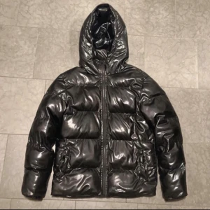 Svart glansig pufferjacka med huva - Säljer en svart pufferjacka med glansig finish och huva. Jackan har dragkedja framtill och breda, vadderade kanaler som ger en riktigt fet look. Perfekt för kalla dagar när du vill hålla stilen och värmen.