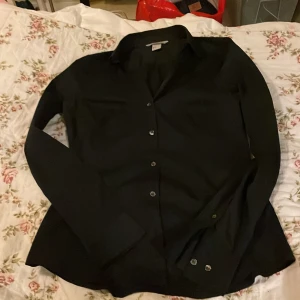 Svart skjorta från H&M, strl 34 - Stilren svart skjorta från H&M i storlek 34. Skjortan har klassisk krage, långa ärmar med dubbla knappar vid manschetten och knäppning framtill. Materialet känns mjukt och lätt, perfekt för en clean look. Passformen är normal och designen är enkel utan mönster.