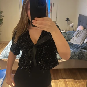 Svart paljett bolerokofta med rosett - Säljer en svart bolerokofta med glittriga paljetter och en stor rosett framtill. Toppen har korta ärmar och är croppad, vilket ger en cool och festlig vibe. Perfekt att styla över en enkel topp eller klänning för att lyfta din look.