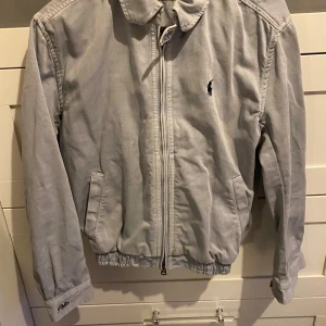 Ljusblå bomberjacka Polo Ralph Lauren XS - Snygg ljusblå/grå bomberjacka från Polo Ralph Lauren i bomull. Klassisk design med dragkedja, krage, två fickor och broderad logga på bröstet. Elastisk nederkant och ärmslut, samt Polo-broderi på ärmen. Perfekt för dig som gillar stilren och tidlös look.