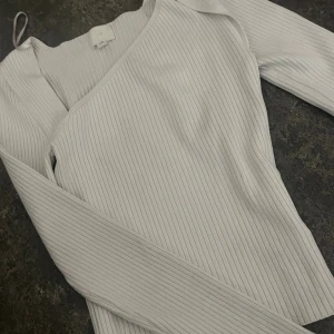 Vit ribbad omlottopp med lång ärm - Säljer en vit omlottopp från H&M med ribbad struktur och lång ärm. Toppen har en snygg asymmetrisk halsringning och är tillverkad i mjuk bomullsmix.