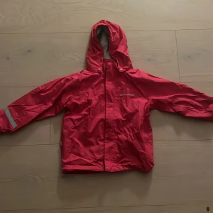 Röd regnjacka från Didriksons - Snygg röd regnjacka från Didriksons med huva och reflexdetalj på ena ärmen. Jackan har dragkedja framtill och är tillverkad i ett vattenavvisande material. Perfekt för regniga dagar och utomhusaktiviteter. Storlek 122