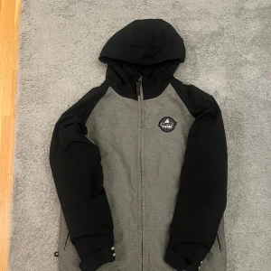 Grå och svart skidjacka från Burton Xs - Storlek 164-169, XS, Barnstorlek XL. Snygg skidjacka från Burton i grått och svart med huva och hel dragkedja. Jackan har svarta ärmar och huva, grå kropp och diskret Burton-logga på bröstet. Perfekt för kyliga dagar och skid åkning. 