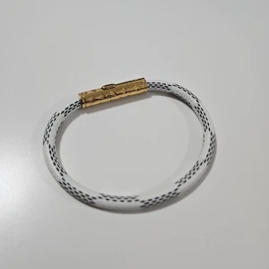 Vitt armband med guldspänne - Snyggt armband med vit bas och svarta detaljer i ett mönster längs bandet. Spännet är i guldton med ingraverade detaljer som ger en lyxig känsla. Perfekt accessoar för att lyfta din outfit.