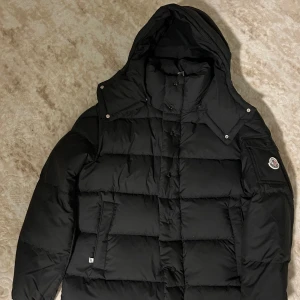 Svart dunjacka från Moncler med huva - Säljer en svart dunjacka från Moncler med avtagbar huva och klassisk logga på ärmen. Jackan är quiltad, har dragkedja och knappar framtill samt praktiska fickor. Perfekt för kalla dagar och riktigt snygg streetstil.