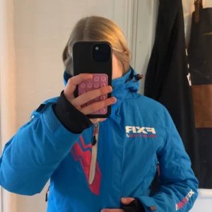 Blå rosa FXR vinterjacka med dragkedjor - Säljer en blå vinterjacka som jag har andvänt en vinter den är från FXR med coola rosa och vita detaljer samt flera dragkedjor. Jackan har huva, praktiska fickor och snyggt mönstrad logga på bröstet. Perfekt för kalla dagar och outdoor-äventyr. Sitter skönt och har sportig look.