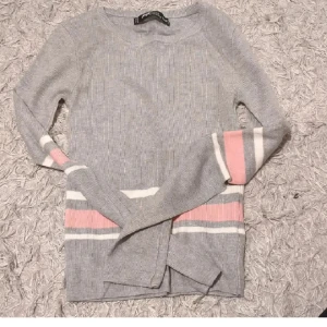 Grå ribbad stickad tröja med ränder - Snygg grå stickad tröja med ribbad struktur. Tröjan har lång ärm och coola ränder i rosa och vitt på ärmar och nedtill. Perfekt för dig som gillar en enkel men ändå trendig look.