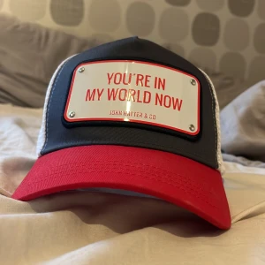 John Hatter & Co röd och vit keps - Snygg truckerkeps från John Hatter & Co med röd böjd skärm, vit mesh på sidor och bak samt mörkgrå front. Metallskylt med texten 'YOU'RE IN MY WORLD NOW' framtill. Justerbar snapback och cool streetstil. Perfekt för att sticka ut.
